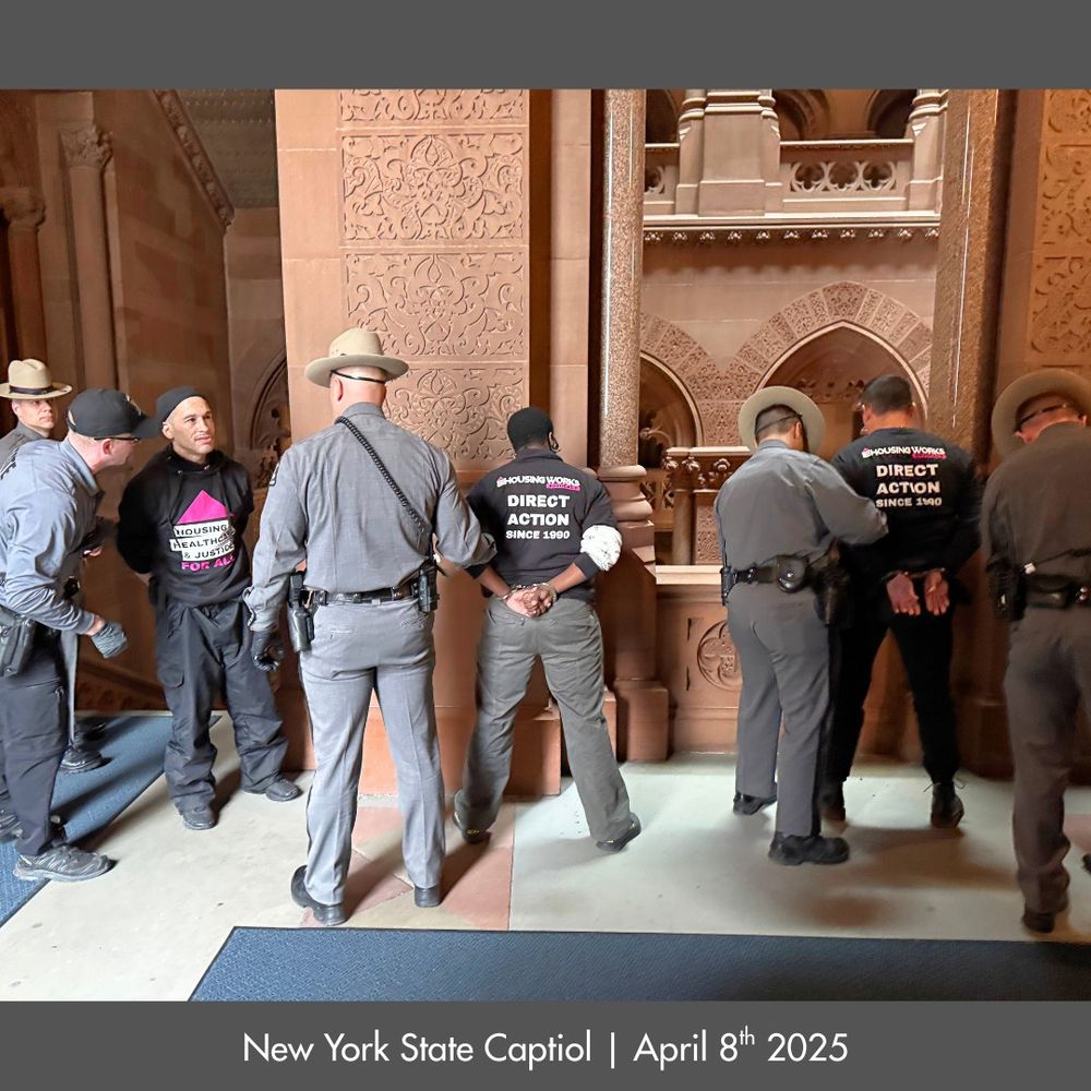 Lined up between five cops, three men stand handcuffed with their hands behind their backs. One has turned to speak to an officer; the front of his shirt reads “Housing Healthcare & Justice For All” with a large pink triangle. The other two are held facing a railing; the backs of their shirts read “Housing Works Advocacy: Direct Action Since 1990”
