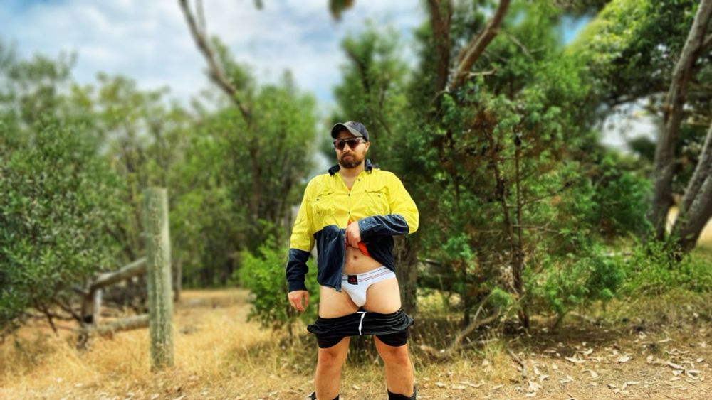 An individual stands confidently in an Australian bush setting, wearing a high-visibility yellow work shirt, sunglasses, and a cap. Their pants are lowered to reveal a white jockstrap, blending rugged masculinity with playful boldness. The dry grass and native trees evoke the essence of the outback.