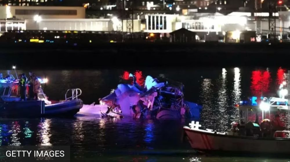 An image from Getty Images, taken of a night time scene on the Potomac river. Two small search vessels illuminate a broken UH-60 Blackhawk helicopter, in the water.  In the background you can see lit up buildings, and lights reflecting off the water.