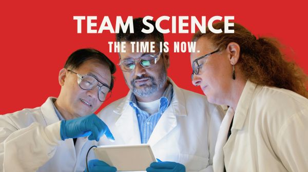 A cutout of three lab scientists looking at an iPad over a red background, with the following text in white, all caps: (line 1; larger) TEAM SCIENCE; and (line 2) THE TIME IS NOW.