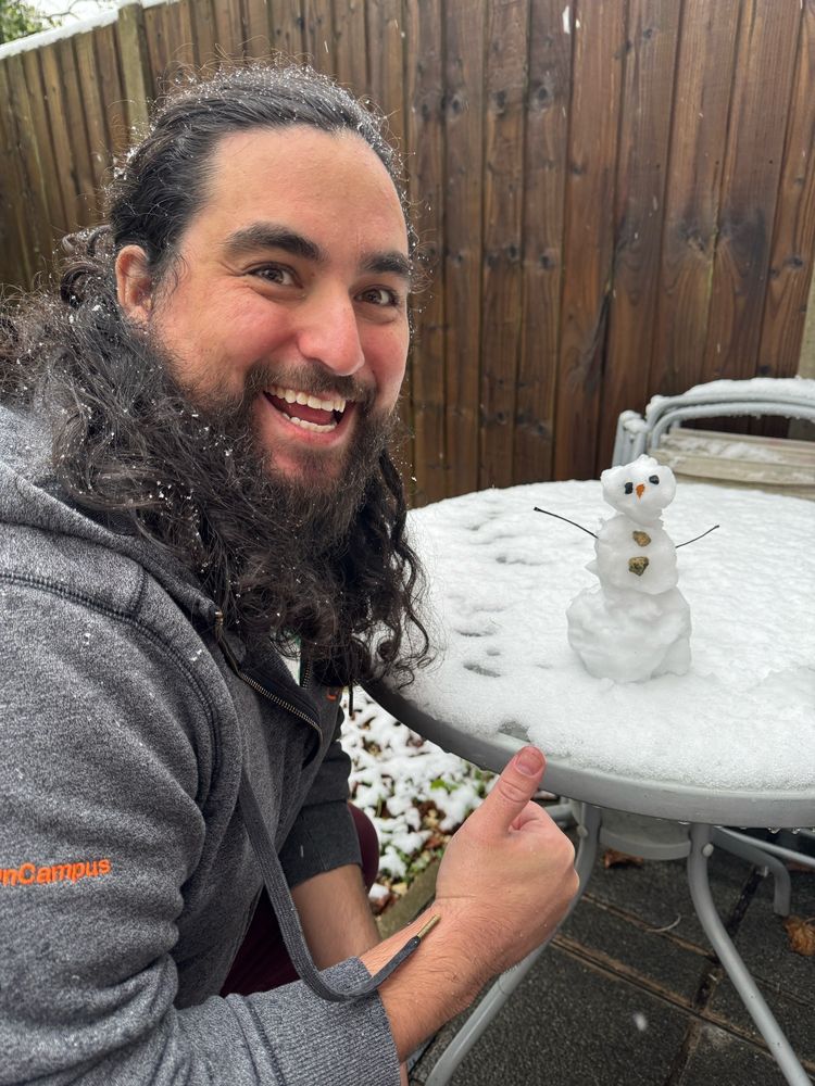 A dashingly handsome man with long hair and a beard giving an approving thumbs up to an adorable little snowman on a table. 