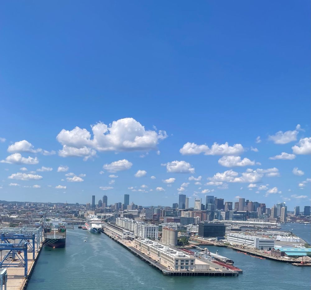 View of Boston Harbor from an airplane landing at Logan. You can see the piers, the skyline, and a beautiful blue sky.