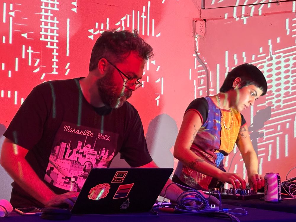 Two digital artists with laptops and various control devices, staring intently at their screens, with a glitchy abstract red and white video projection over them both