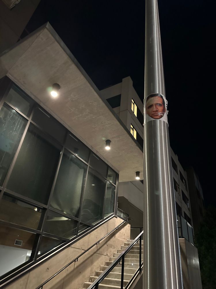 A post beside a brutalist building. High up the post there is a picture of Nicolas Cage