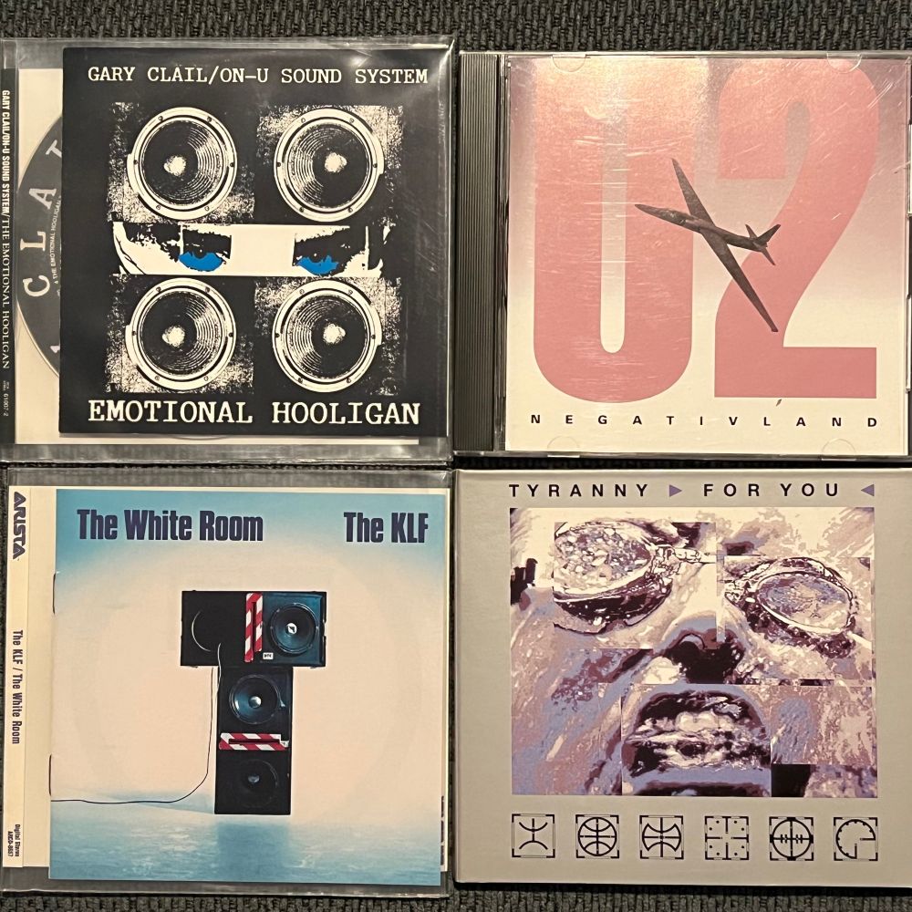 Four CDs arranged in a 2x2 square. Clockwise from top-left corner: “The Emotional Hooligan” by Gary Clail/On-U Sound System, “U2” by Negativland, “Tyranny (For You)” by Front 242, and the U.S. version of “The White Room” by The KLF. 