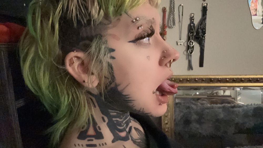 Side profile of heavily tattooed individual with a lime green mullet sticking out their split tongue