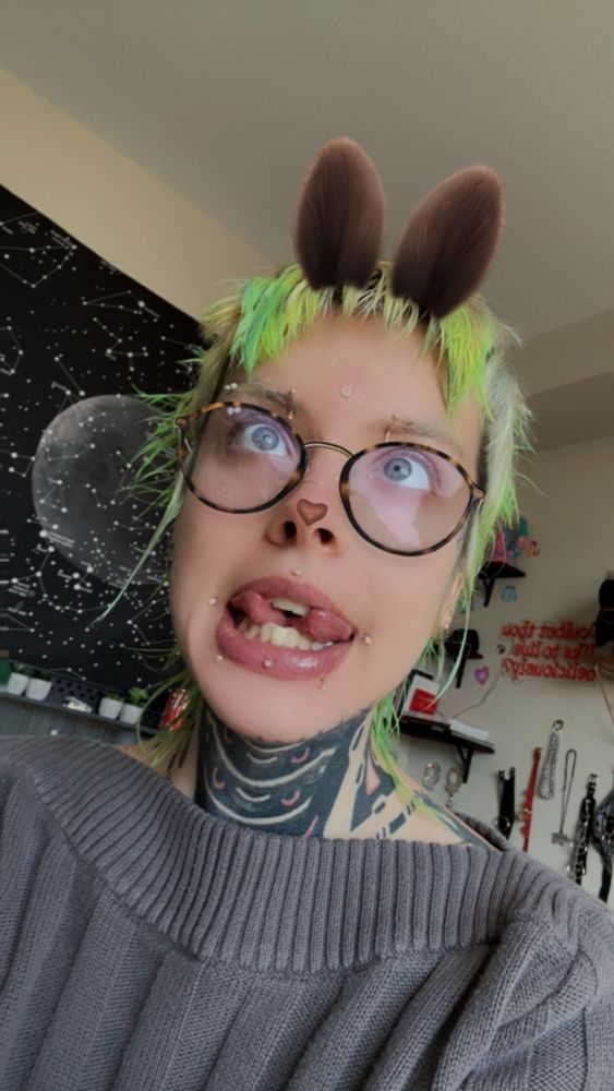 Short green haired mullet boy makes a silly face, split tongue separated and showcasing a new dermal I no the center of their forehead