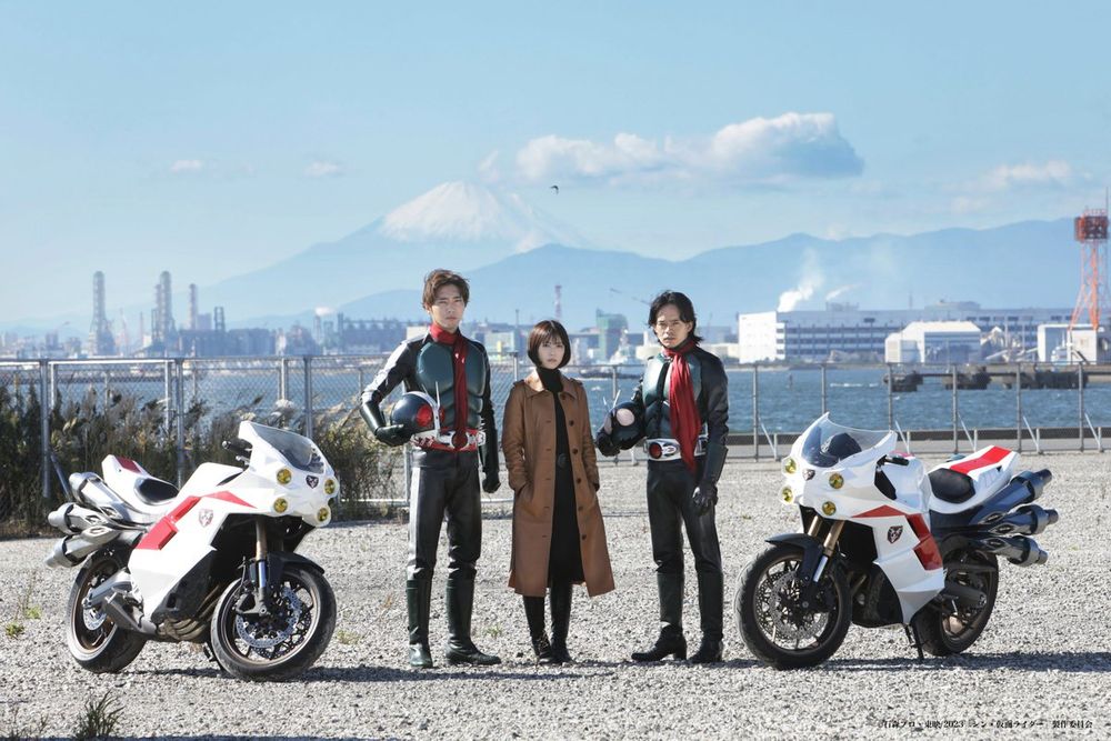 Hayato Ichimonjo, Ruriko Midorikawa, and Takeshi Hongo from Shin Kamen Rider flanked by their motorcycles, Cyclone, at a city with a bay area behind them. 