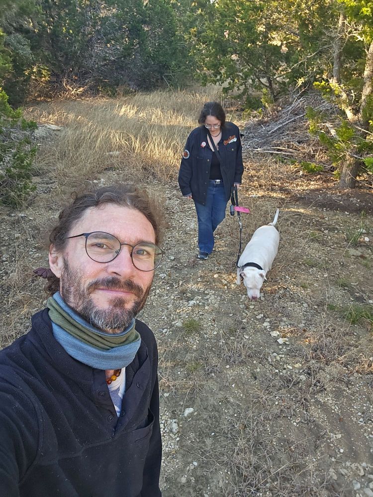 Bearded man with a neck scarf taking a selfie with a woman dressed in a denim jacket walking a white dog on a leash in the background. 