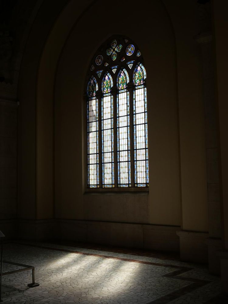 A gothic like stained glass window. The light goes through it and is reflected over the floor, the space is dark creating a contrasting effect.
