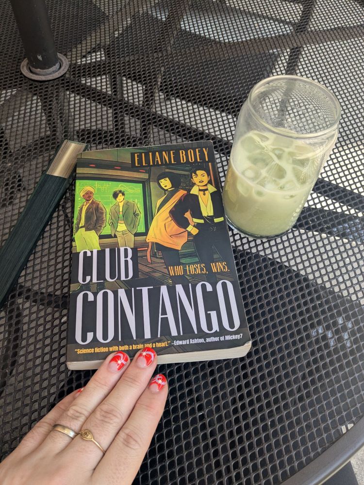 Club Contango by Eliane Boey on a table between a hand fan and a matcha latte, all in similar greens and golds. (Part of my hand is there too.)