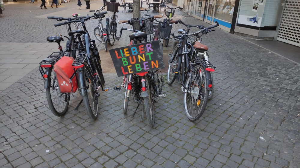 Ein paar Fahrräder stehen nebeneinander. An einem davon ist ein großes, schwarzes Schild angebracht, auf dem in großen bunten Letter LIEBER BUNT LEBEN geschrieben steht.