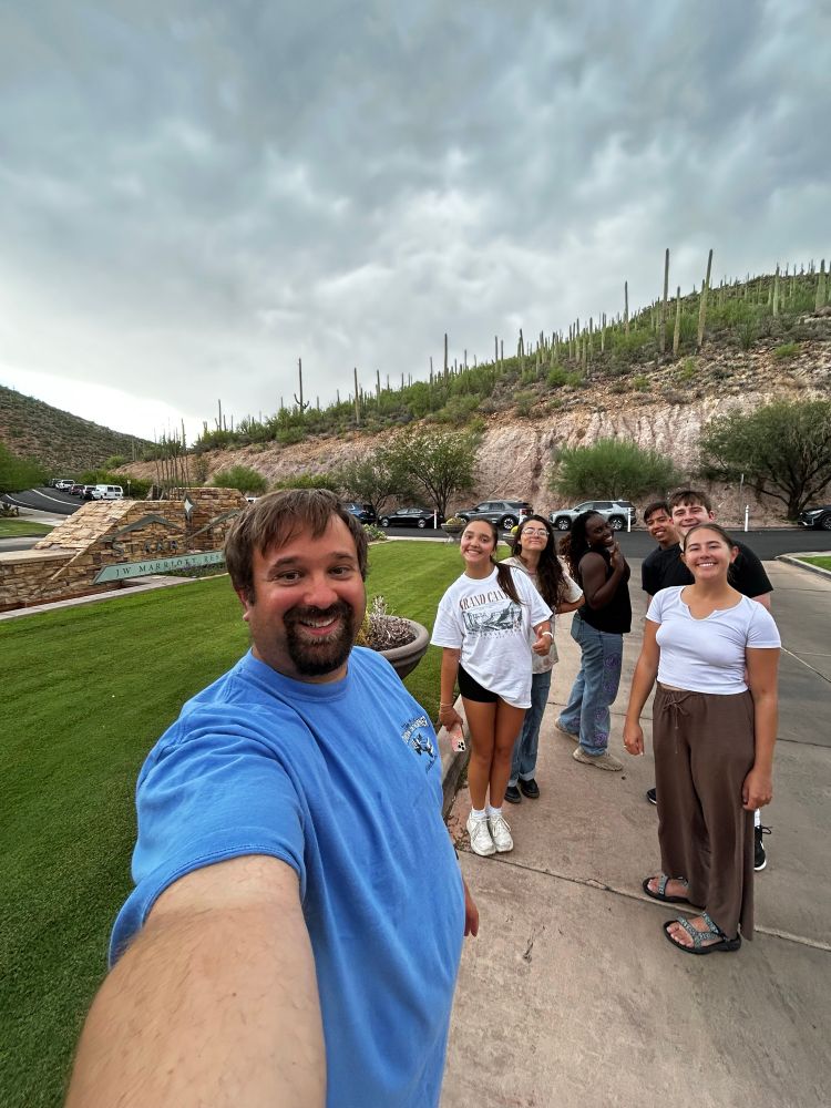 Selfie of the lab in front of the entrance to Starr Resort in Tuscon, the location of Botany 2026. Saguaros in the background!