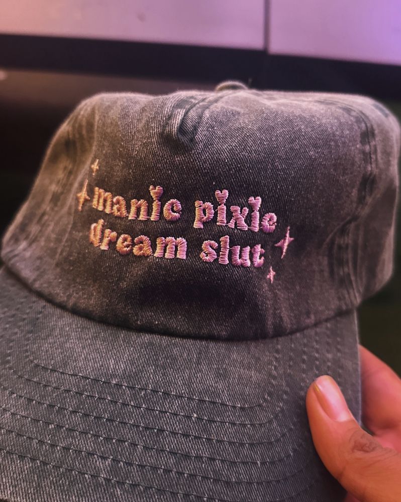a picture of a green baseball cap which reads “manic pixie dream slut”