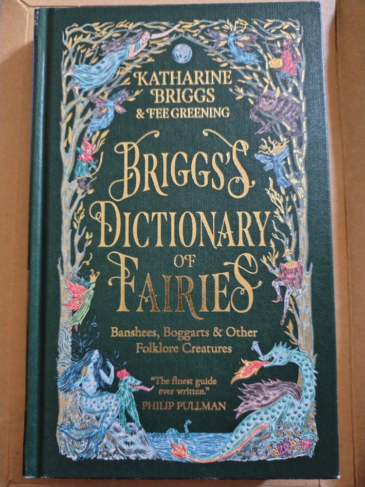 The front cover of Briggs's Dictionary of Fairies by Katharine Briggs & Fee Greening. The background is forest green, the text of the title and author is gold. On either side is a tree in gold and grey, the branches meet forming an arch at the top. In the branches are fairies and other magical creatures, decked in red, blue, yellow and green. At the bottom is a lake with what could be Nessie in it. On one side of it is a blue and red dragon breathing fire, on the other side is a mermaid combing her long dark hair.
