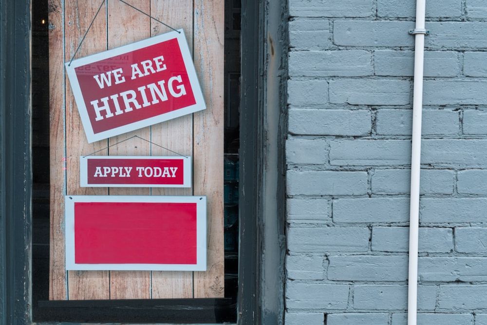 A shopfront window displays two red-and-white signs reading “We Are Hiring” and “Apply Today.” The empty bottom panel underscores how opportunities are shrinking, symbolizing the gap between visible job ads and the reality of scarce, unstable work for young Canadians in 2025.