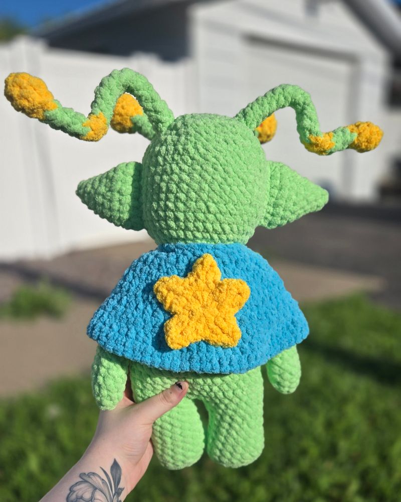 back shot of crocheted green ailen. his blue cape has a large yellow star in the middle
