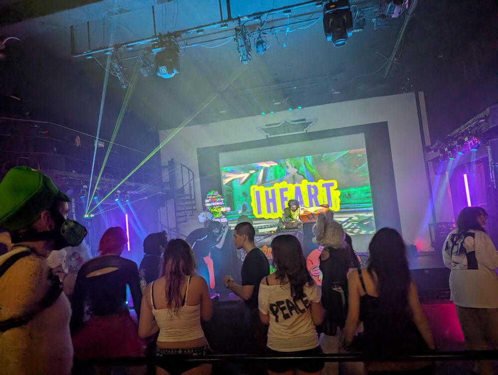 A picture looking out at the crowd of a rave. There are lasers and projection spotlights beaming across the foggy atmosphere. Several people are wearing fursuit heads. The stage has a large projection screen which shows "IHEART" in big neon lettering. 