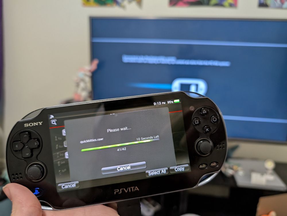 A PlayStation Vita being held in front of a TV with a PS3 displaying a connection message. The PlayStation Vita is transferring music files to its memory card from the PS3. 