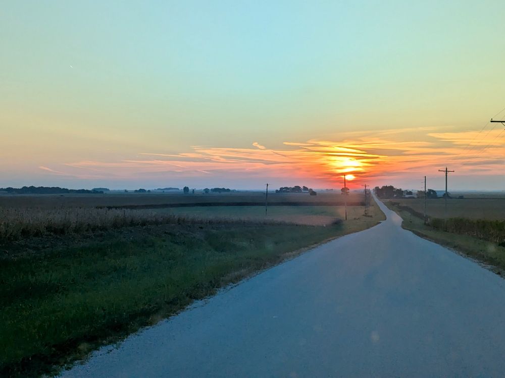 A sunrise behind distant clouds warmly lights a road narrowing into the distance at center right of the frame. Some fields are to the left, and the sky is a pale blue in the upper third of the image. 