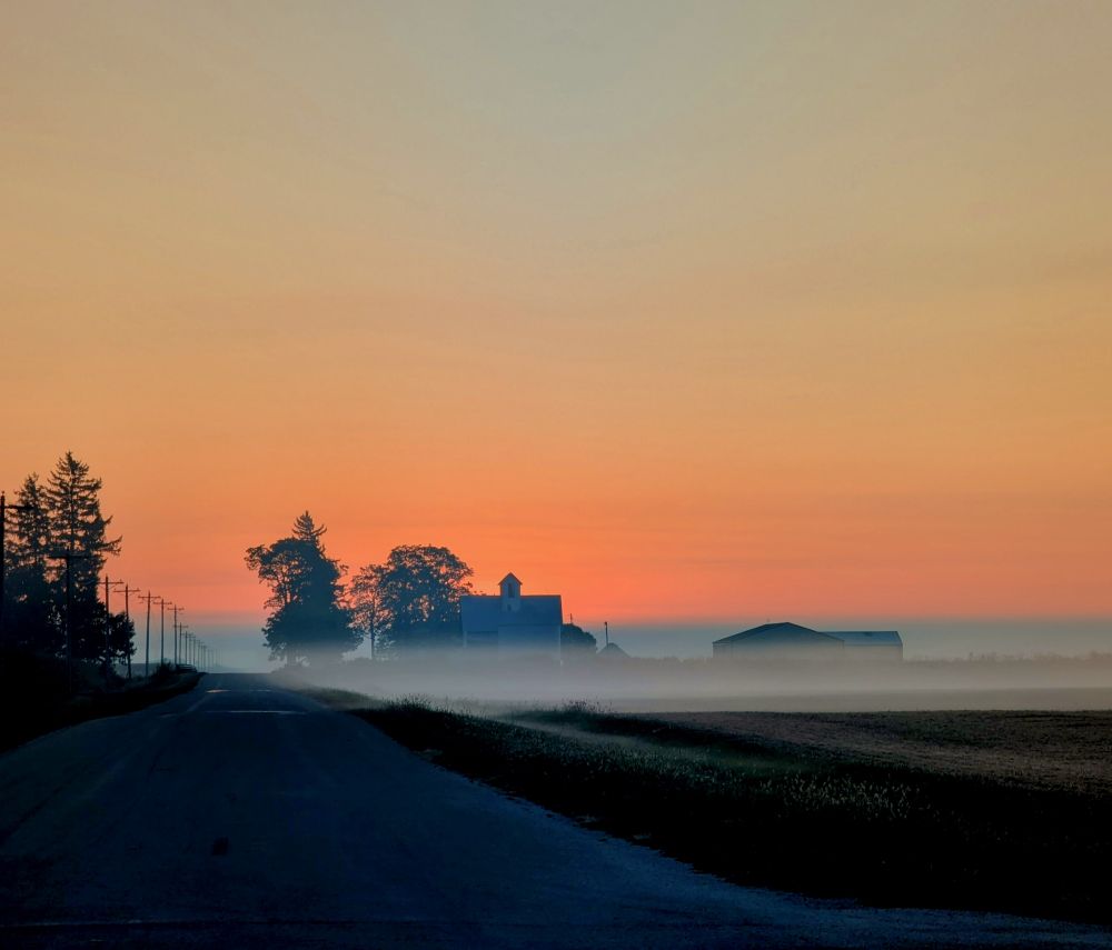 An image of a rural area at sunrise. The foreground shows a darkly lit road at left narrowing into the distance. At the horizon in the center is a far off building, likely a barn and another one to its right hidden partly by morning fog. The sky is warm and orange at the horizon and then slowly fades to a pale bluish-grey at the top. The scene is peaceful and serene.