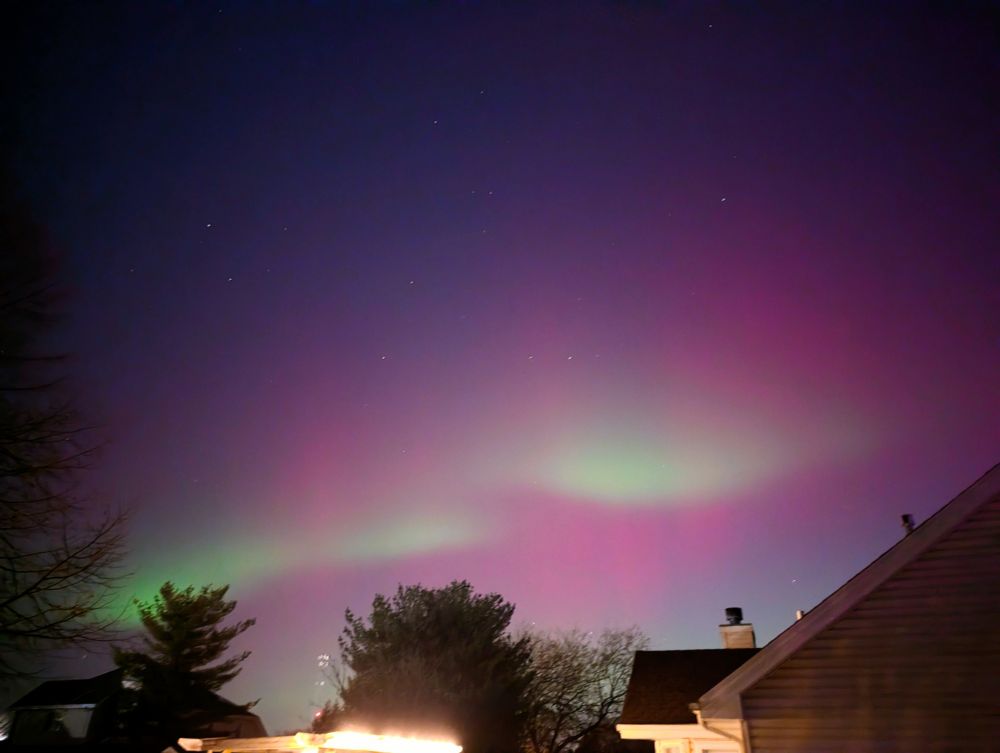 A green Aurora goes from lower left towards middle right with pink Aurora above and below it. There is a house roof to the lower right and some trees at the bottom.