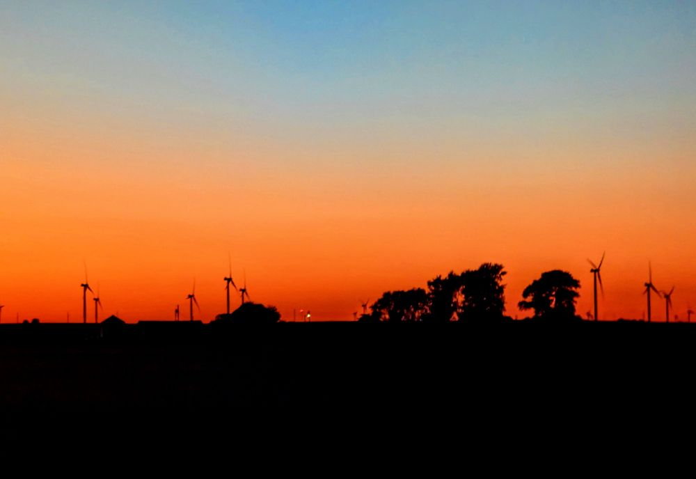 A dusk scene. The horizon is black. There are some wind turbines and trees along the horizon. A deep orange fades slowly into a lighter orange upward, then turns light blue into a deeper blue at the top.