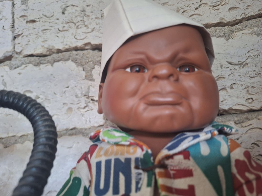 Plastic black baby wearing a paper hat lookin in an interesting way. He is like questioning you. Sits before a wall. Wears a jacket with things typed on it in a stylized way.