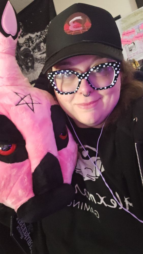 A photo of Beth in a hat, black and white polka dot glasses, and a Alexmaria530 gaming shirt, while hugging a pink baphomet head. 