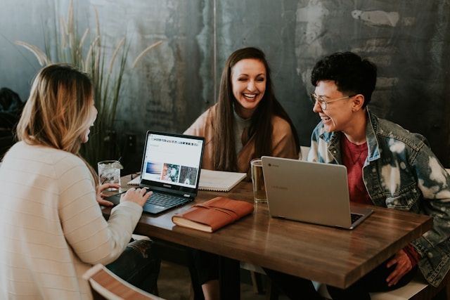 Photo by Brooke Cagle on Unsplash. Three young professionals sharing a laugh while working together at a wooden table with laptops and notebooks. They're in a modern industrial-style space with concrete walls.