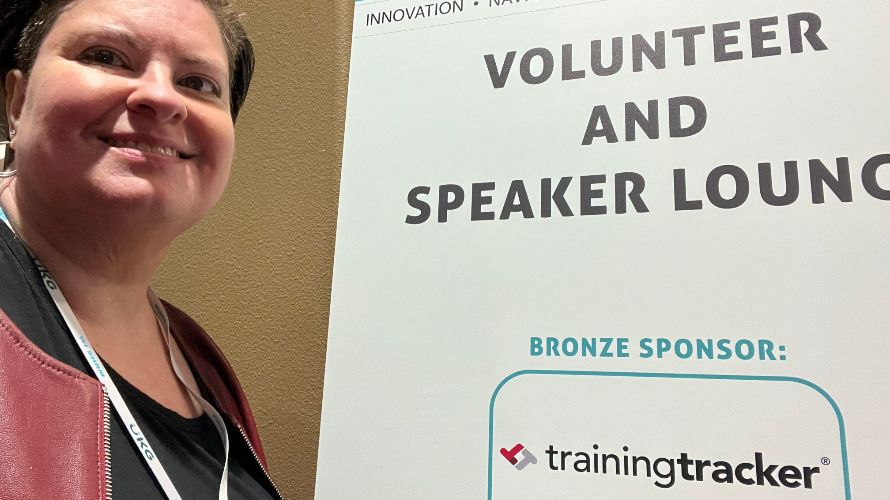 Professional woman with short dark hair wearing a lanyard and burgundy jacket smiles at the camera while standing next to a conference sign reading "VOLUNTEER AND SPEAKER LOUNGE" with "BRONZE SPONSOR: trainingtracker" displayed below. The setting appears to be at an innovation or professional development event, with a beige wall visible in the background.