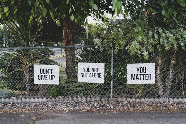 A chain-link fence in a green area with three white signs attached to it. The signs read, from left to right: "DON'T GIVE UP," "YOU ARE NOT ALONE," and "YOU MATTER." Lush trees and foliage are visible in the background.
