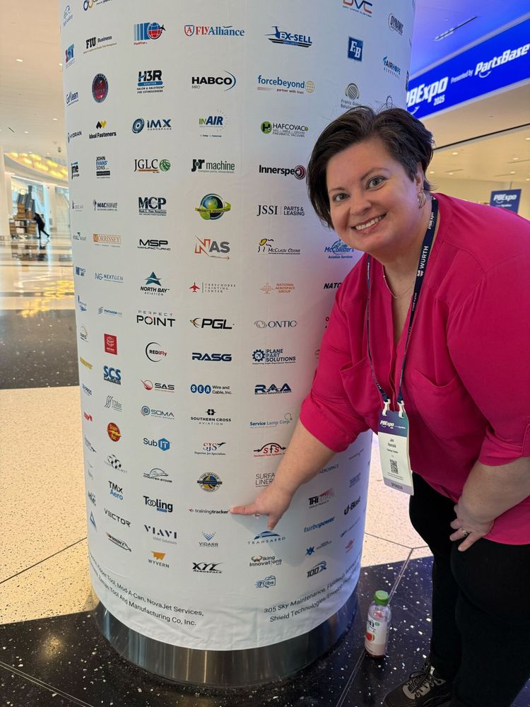 A woman in a bright pink top at a conference smiling and pointing to the Training Tracker logo on a cylindrical sponsor display pillar covered with various company logos and sponsors of the PBexpo event.