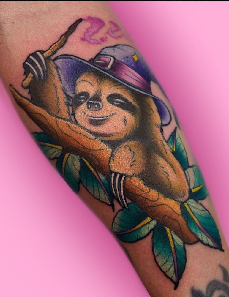 A colorful sloth tattoo. The sloth is wearing a purple wizard hat with a strap and stars, casting a snooze spell with a stick wand. The sloth is holding onto a branch with blueish green leaves.