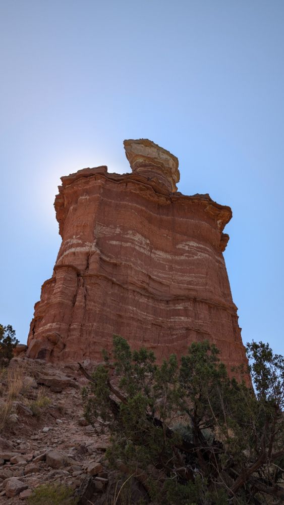 At the base of Lighthouse Rock, looking up at it with the sun beaming from behind the left side of the pillar. Tan and white streaks layer amongst the red rock.