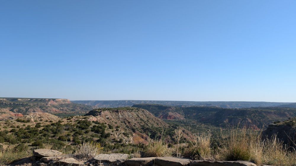 View of Palo Duro canyon from the road along the rim. Canyon formations are banded together with layers of reds, yellows, whites, greys, and browns, speckled and accented with the greens of arid grasses, shrubs, and other plantlife.
