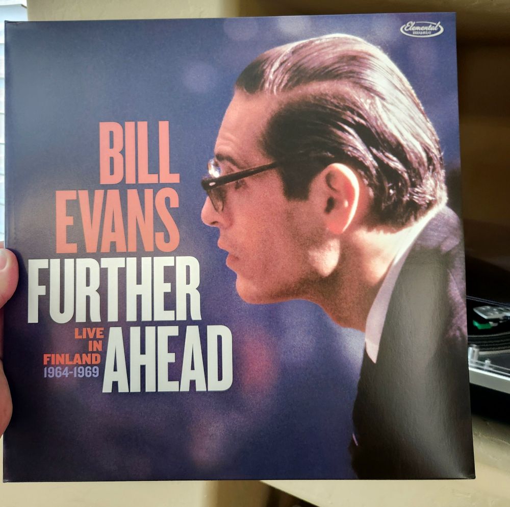 Foreground is record cover of Bill Evans Further Ahead, Live in Finland 1964-1969. Cover depicts Evans bespectacled profile, looking left, with slicked back hair, wearing a dark suit with white shirt and dark tie. 
In the background of this image is a turntable spinning with this album. 