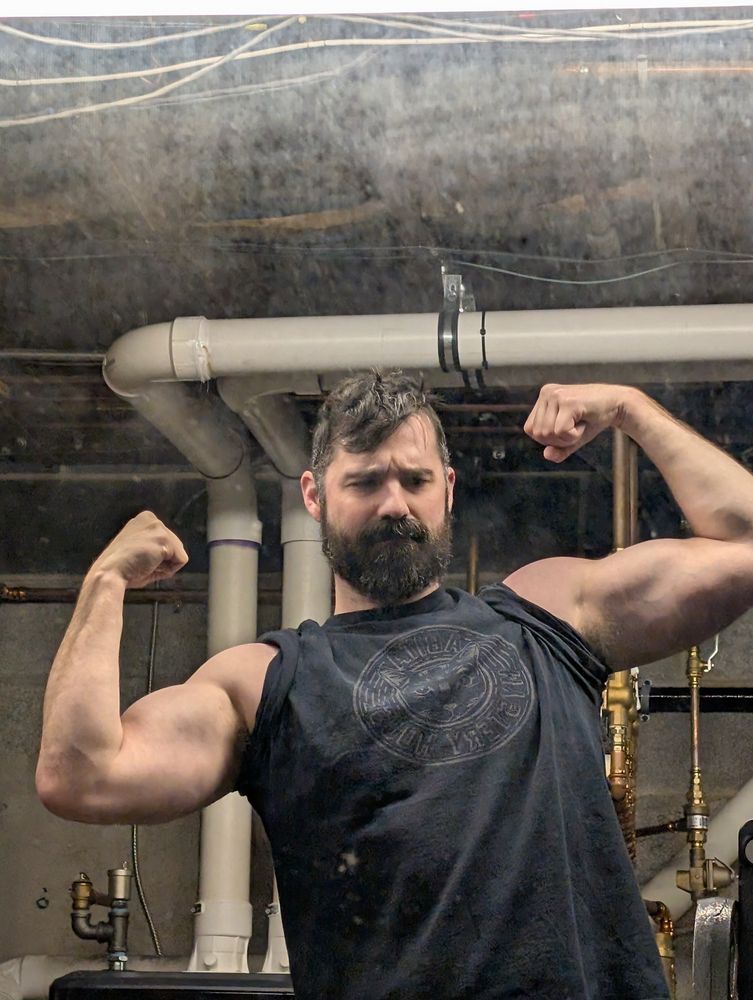 Image of a bearded man with dark hair flexing both biceps. 