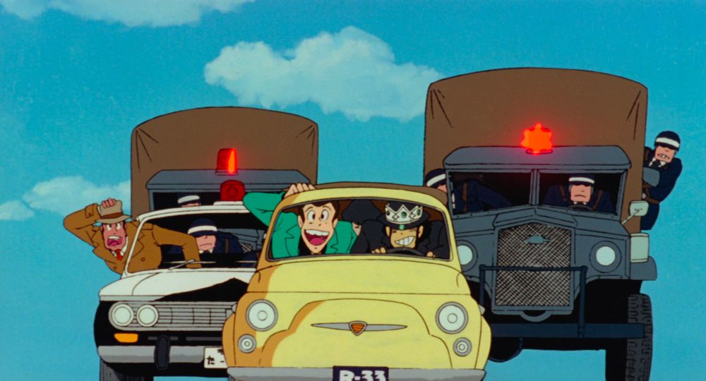 Three handsome men in a Fiat 500, being chased by a man with a prominent chin in a police car and two trucks also full of policemen. One of the handsome men is wearing a crown over his fedora.