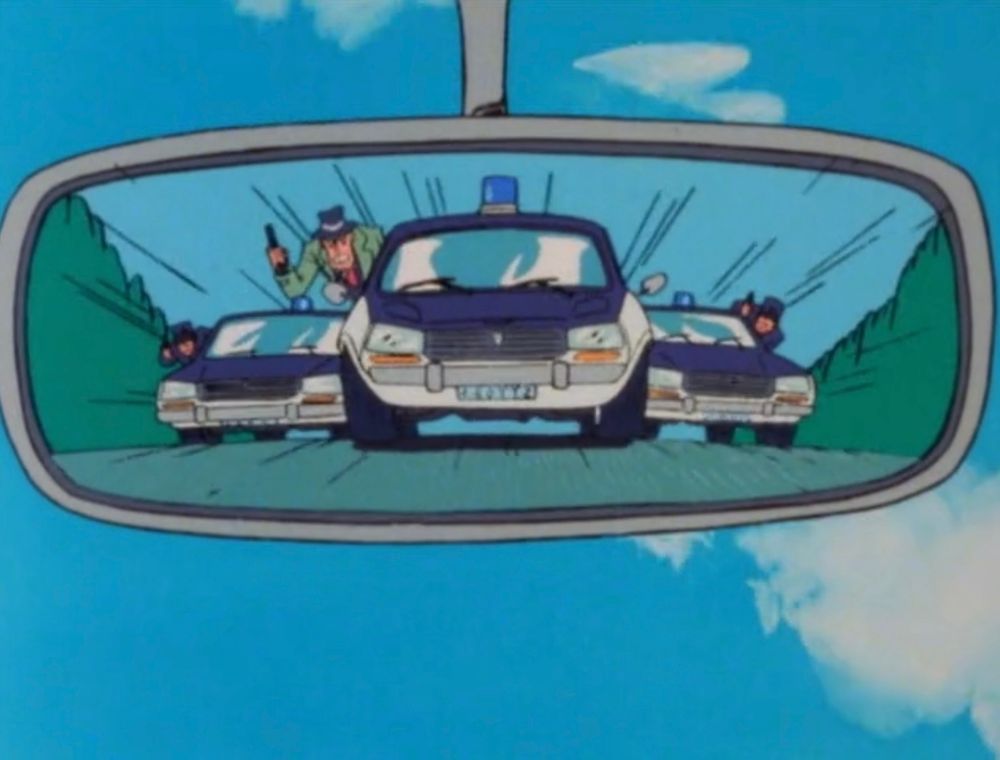 The rearview mirror of a car featuring a view of 3 cop cars. The middle car has a man with a prominent chin and green trenchcoat sticks out of one window, holding a gun.