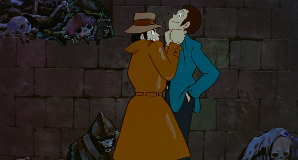 A man in a trenchcoat and hat lifts a handsome man with sideburns, wearing a green jacket, up off the ground. The background is a dull and dingy dungeon, with skeletons.