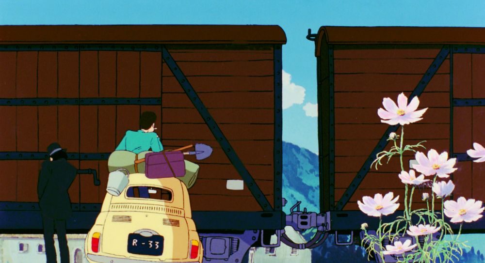 Two handsome men in front of a train in their Fiat 500. The one in the green jacket is looking out the roof of the car. The one in the black suit and hat is taking a whiz on the roadside.
