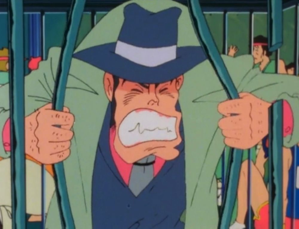 A man with a prominent chin and wearing a mint green trenchcoat pulls apart the bars of the cage he’s been trapped in. His teeth are clenched and he looks angry.