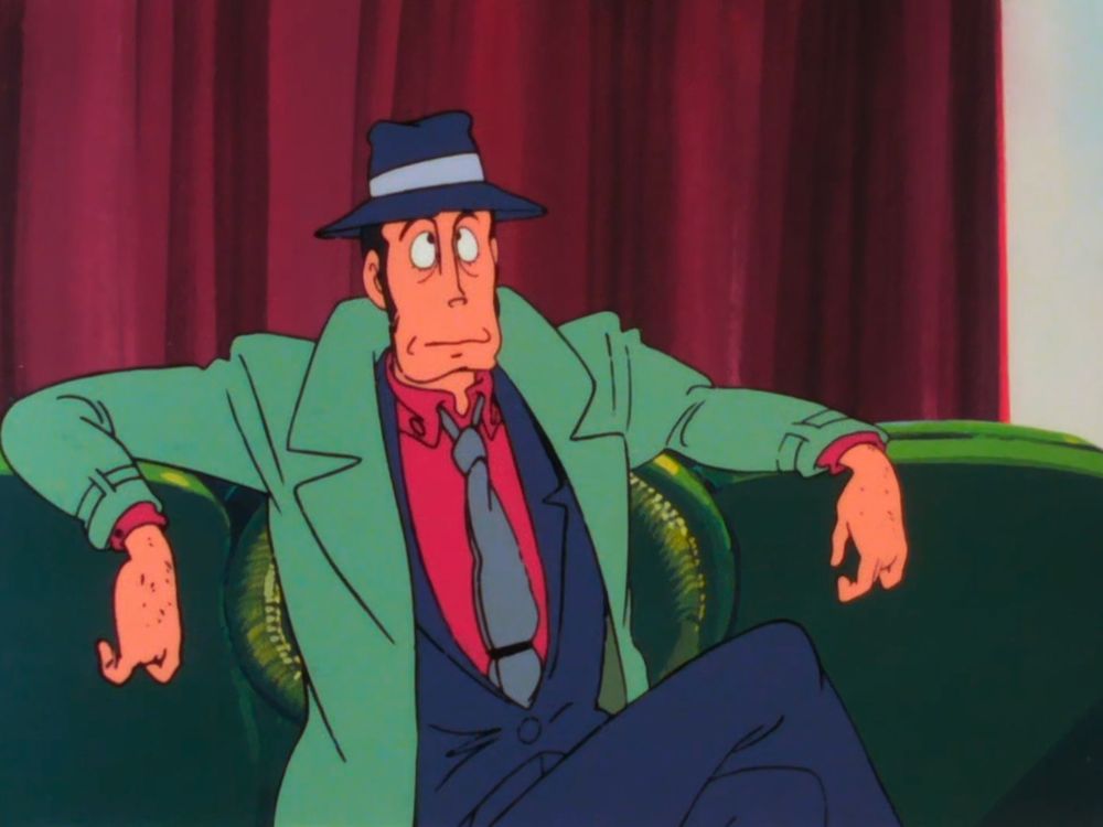 A man with a prominent chin and a green trenchcoat sits on a sofa.