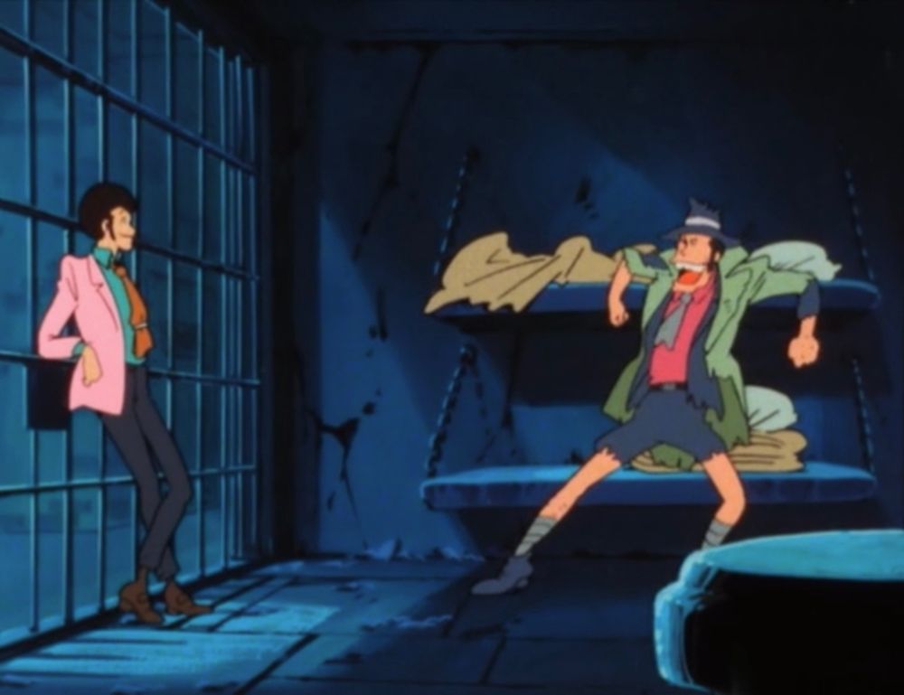 A handsome man in a pink jacket stands in a jail cell with a man with a prominent chin and ripped up clothing.