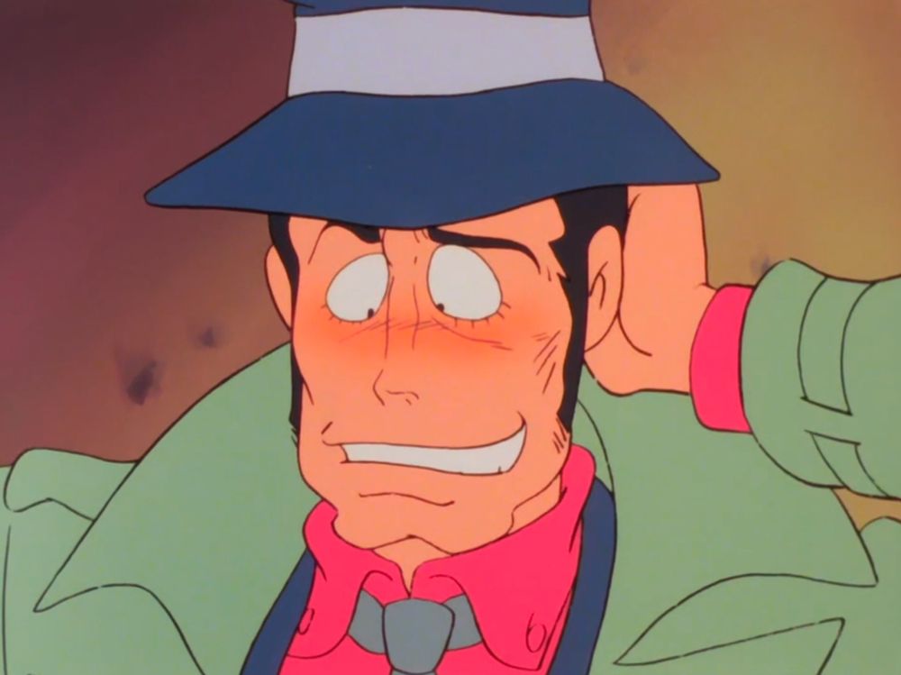 A man with a prominent chin and a green trenchcoat scratches hte back of his head, looking down at the floor and blushing
