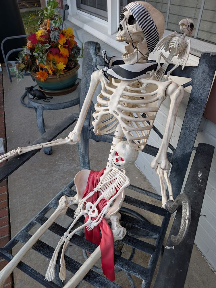 life size pirate skeleton, complete with sashes, headband, eye patch, hook hand and skeleton parrot on his shoulder, sitting in a chair with a toddler sized skeleton in his lap