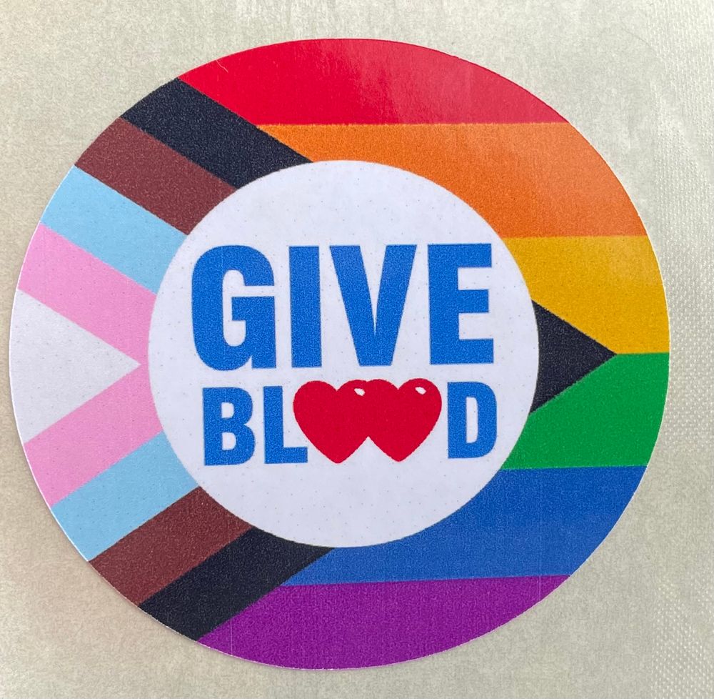 A round sticker displaying the Progress Pride flag with the text “GIVE BLOOD” in the centre, where the double “O” is replaced with red heart icons
