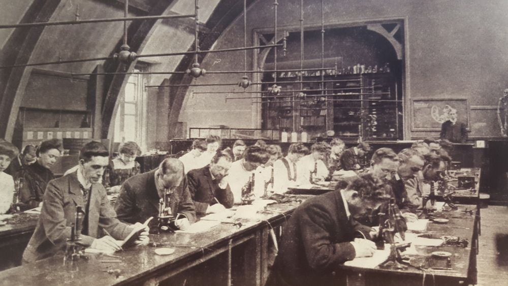 A black and white photo of a biology lab in the early 20th century. Men and women are working at long benches.
