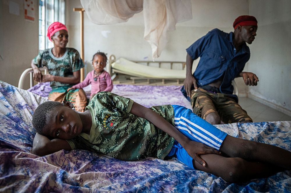 A sick boy lies on a bed in a health center, looking into the camera. His parents and younger sister are sitting on the bed behind him. 
